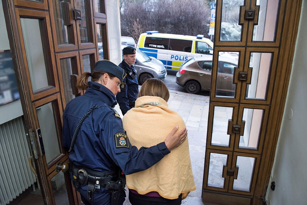 Polis håller om en person inlindad i en filt i en dörröppning. En polis står utanför dörröppningen och väntar vid en parkerad polisbil.