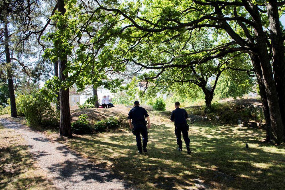 Två poliser går mot två personer som sitter på en bänk. Det är ett grönområde, med träd och man ser poliserna bakifrån.