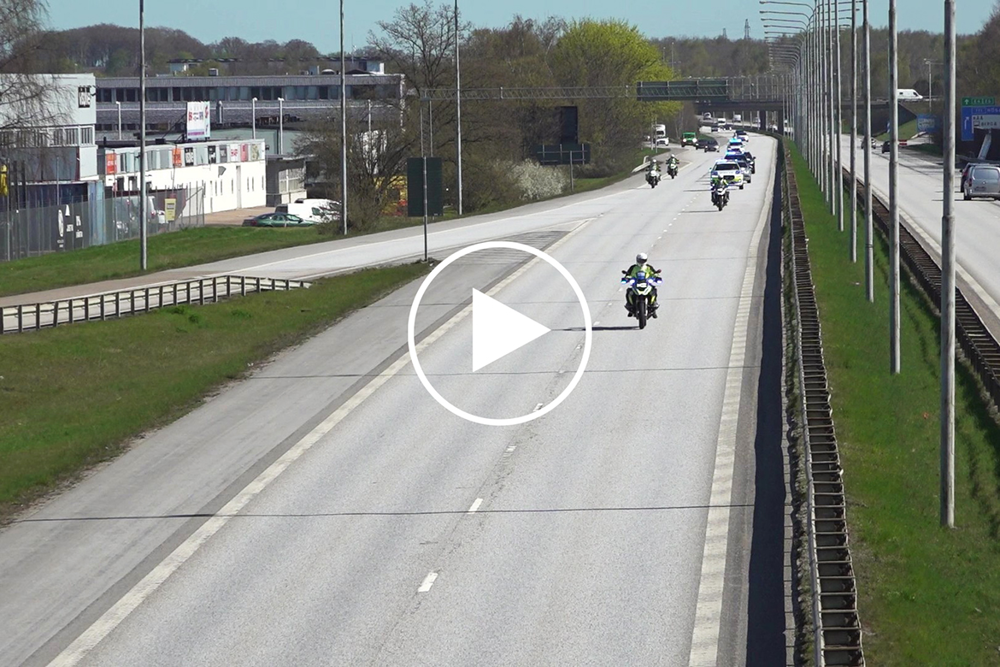 Polismotorcyklar och polisbilar i eskortkörning. Grafisk playknapp visar att detta är en film.