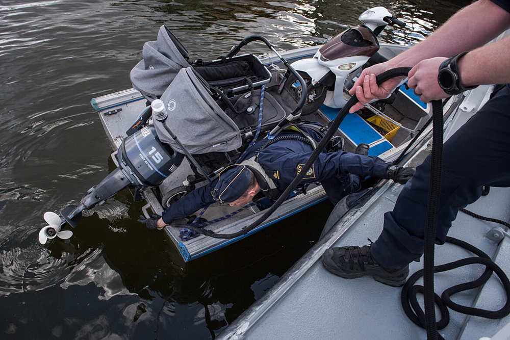 Bilden föreställer två poliser som utreder båtstöld. Den ena polisen befinner sig på båten och sträcker sig mot motorn för att undersöka. Den andra polisen håller i ett rep som båten är förtöjd med.  