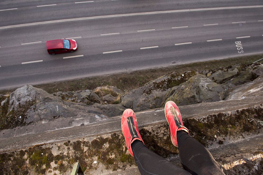 Ett par fötter i röda gympaskor. Nedanför avsatsen åker en bil. vid en klippavsats