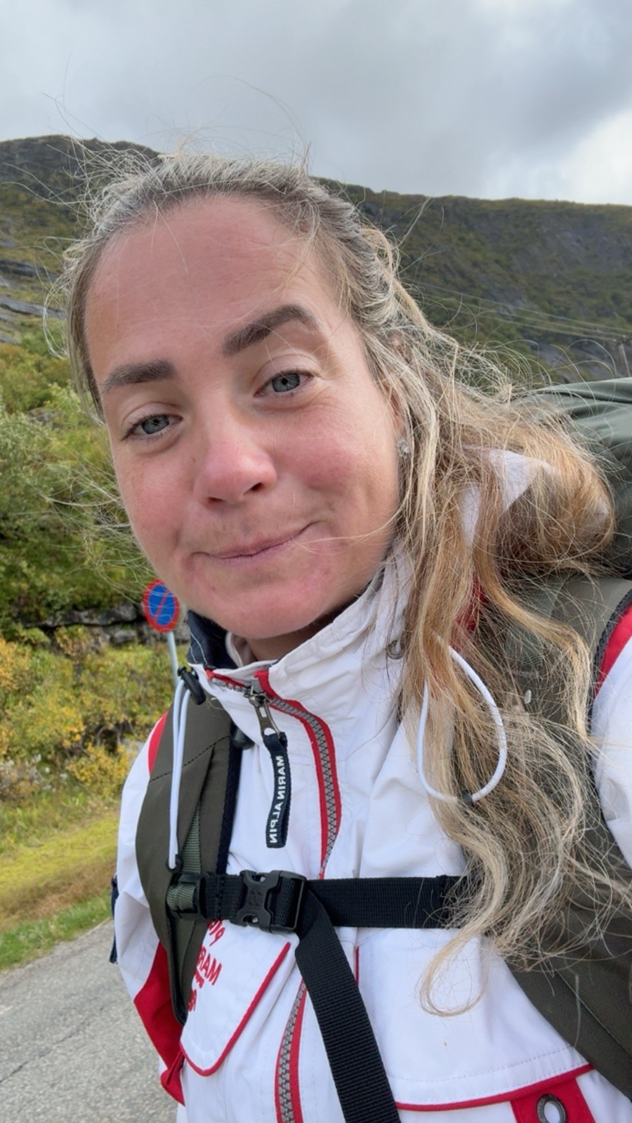 Kvinna med blont hår i en tofs står framför berg och grönska iklädd vit jacka med röda detaljer.