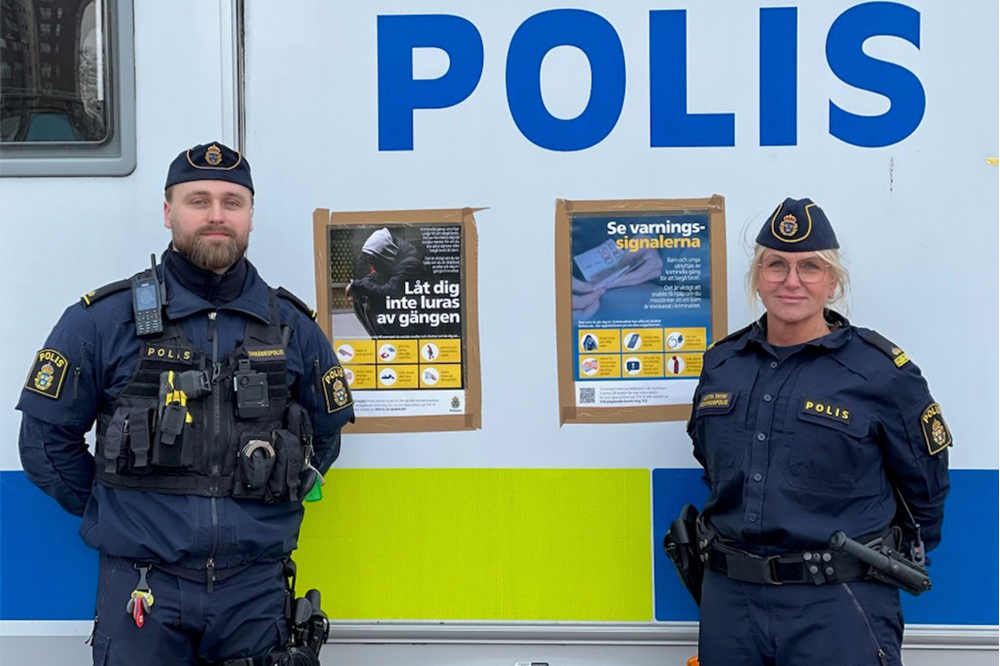 Poliseran Ludvig Bröms och Lotta Thyni står framför en polisbil. I bakgrunden syns affischerna för att motverka gängrekrytering.
