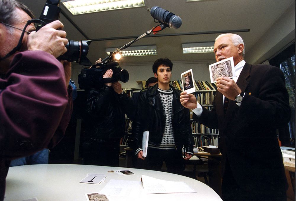 Man på presskonferens håller upp två fotografier framför journalister som har kameror och mikrofoner riktade mot mannen.