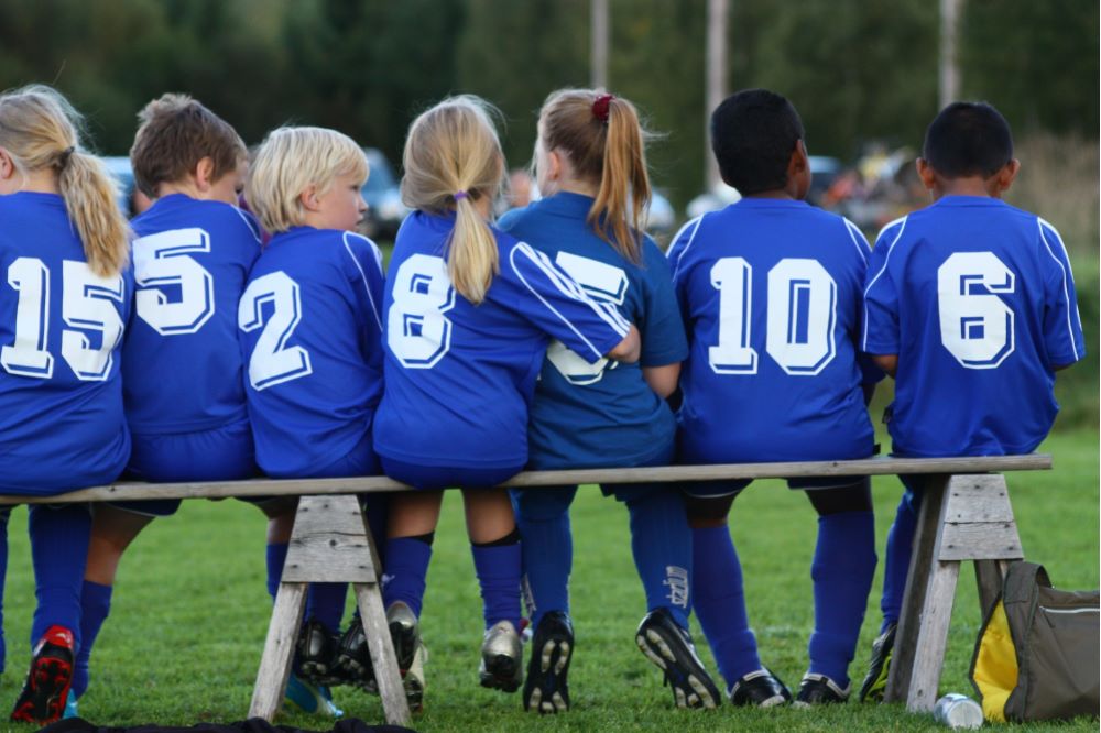 Fyra barn i blå lagtröjor med siffror på ryggen sitter på en bänk.
