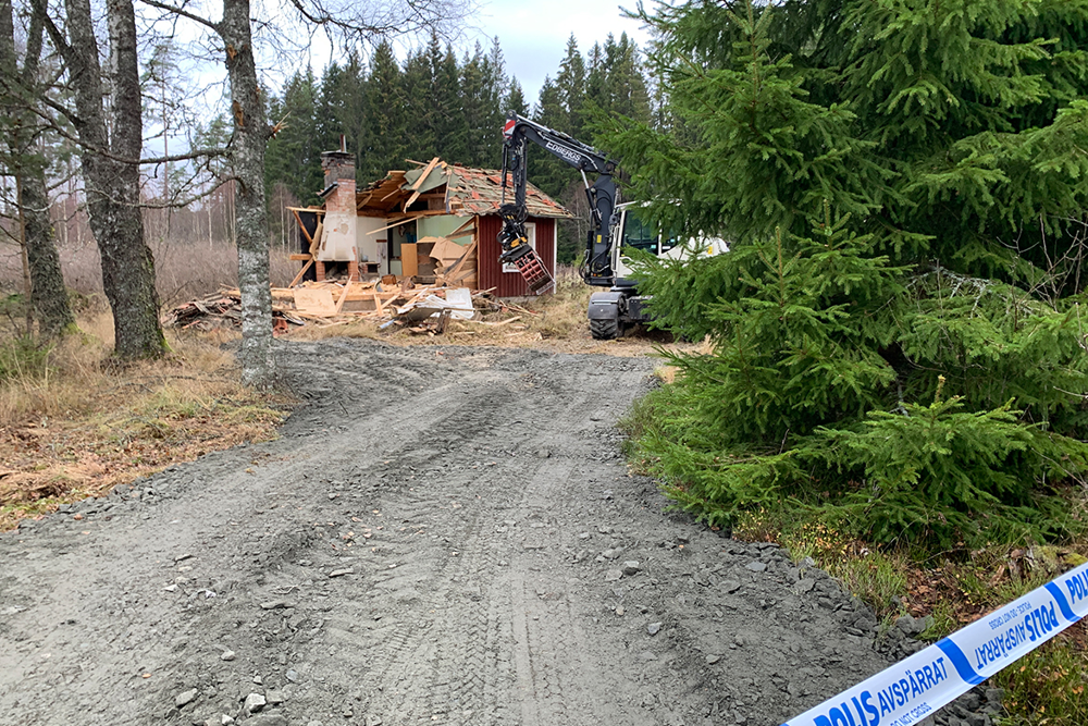 Halvt riven stuga i skogsbryn, med grusväg i förgrunden. Och polisavspärrningsband i nedre kanten av bildens högerkant..