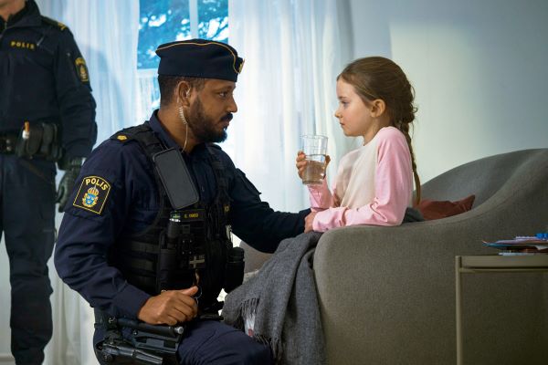 En polis sitter på huk bredvid ett barn som sitter i en fåtölj och dricker ur ett glas. En polis står vid en vägg i bakgrunden.