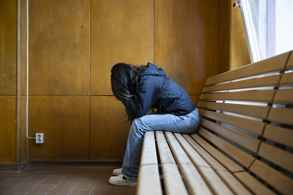 En ung tjej som sitter framåtlutad på en träbänk med huvudet uppgivet placerat i händerna. 