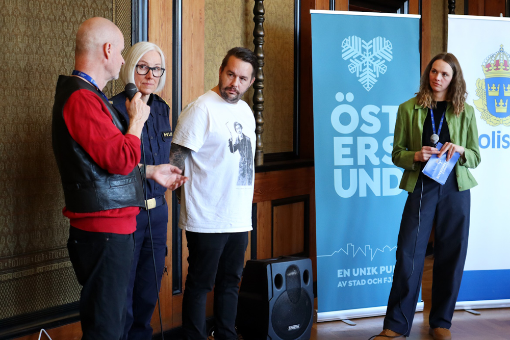 Fyra personer står uppradade bredvid varandra. Mannen till vänster håller i en mikrofon, de andras blickar är vända mot honom. Kvinnan näst längst till vänster har polisuniform på sig, de övriga har civila kläder. I bakgrunden står två roll-ups med texterna Östersund respektive Polisen på sig.