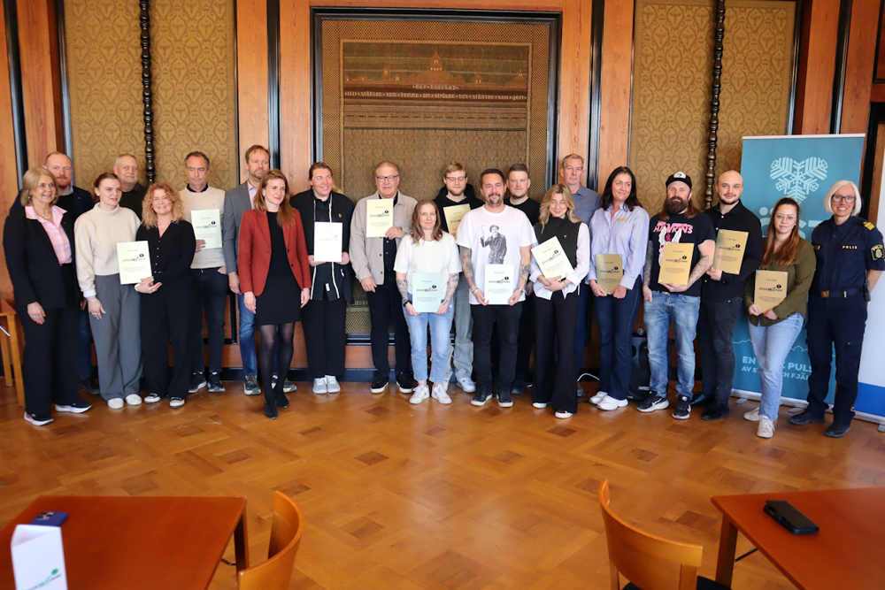 En stor samling av människor står uppradade framför en vägg. Vissa av personer håller i ett intyg i formen av ett papper. Många av personer är glada och ler.