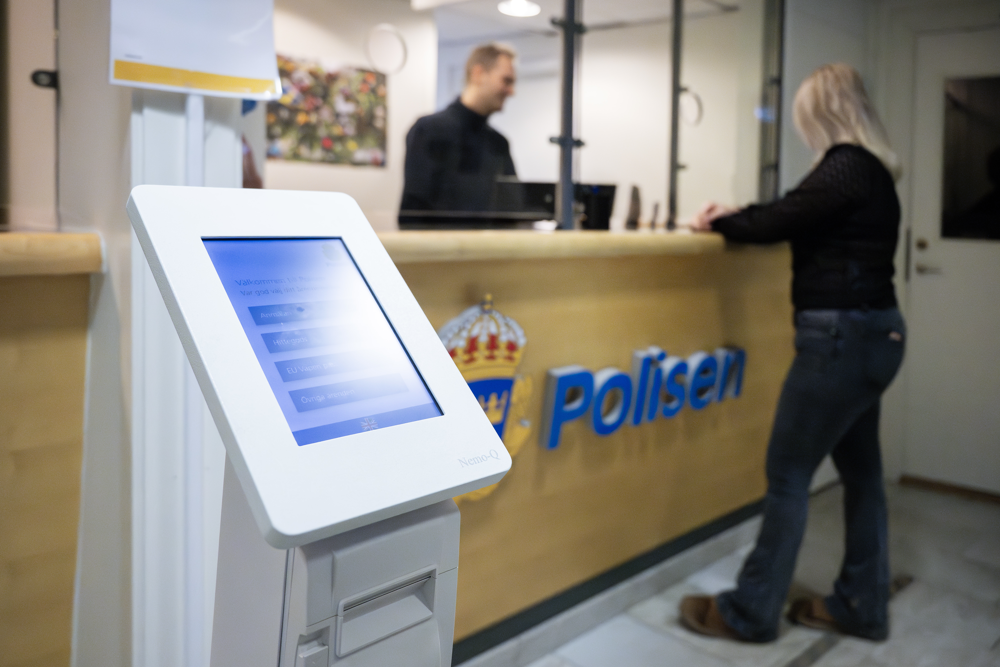 En skärm syns i förgrunden. Bakom den syns en reception och två personer.