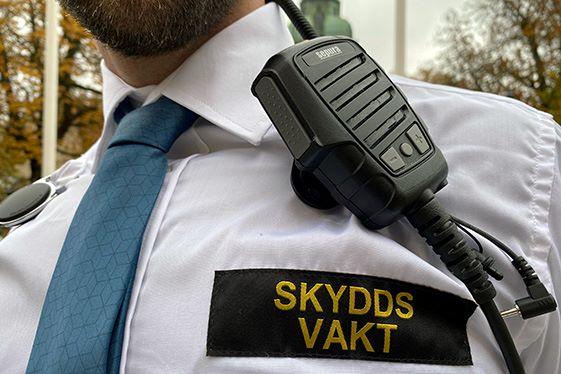 Skyddsvakt med emblem med texten skyddsvakt på en vit skjorta. En kommunikationsradio hänger på skjortan där ovanför .