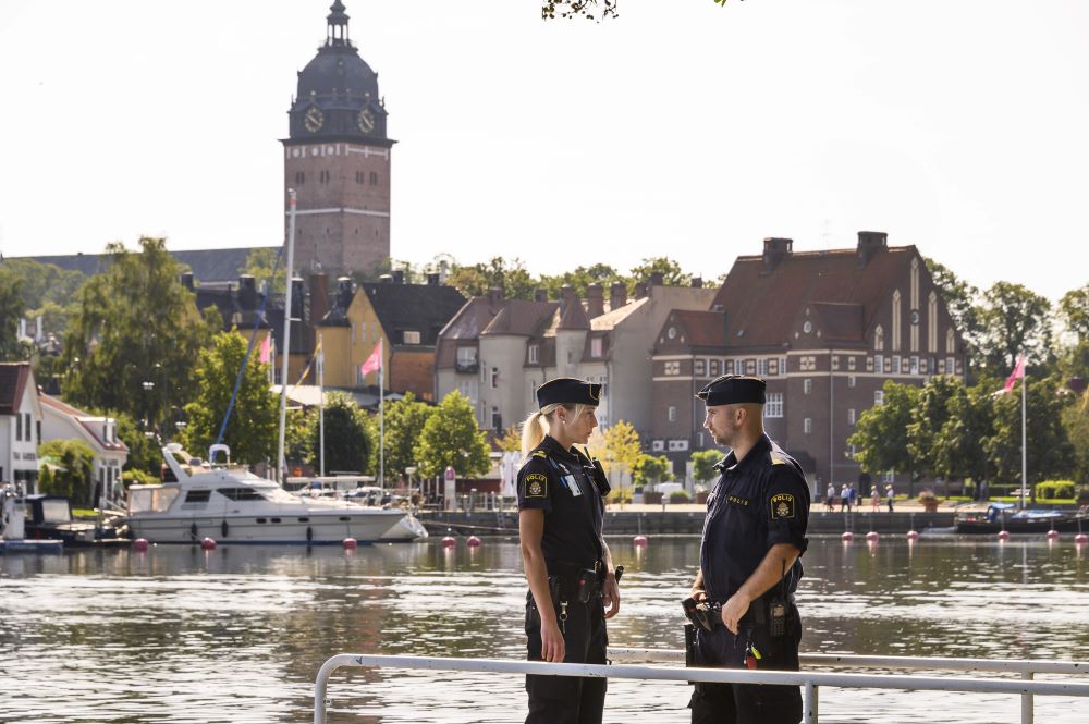 Två poliser, en man och en kvinna, står vända mot varandra och pratar med vatten och stadssilhuett i bakgrunden.