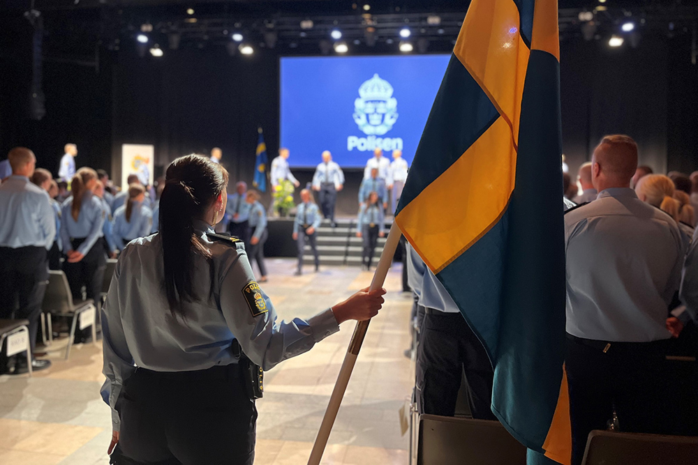 Från avslutningsceremonin: en polis står uppställd med Sveriges flagga, i bakgrunden syns en scen och de nya poliserna.