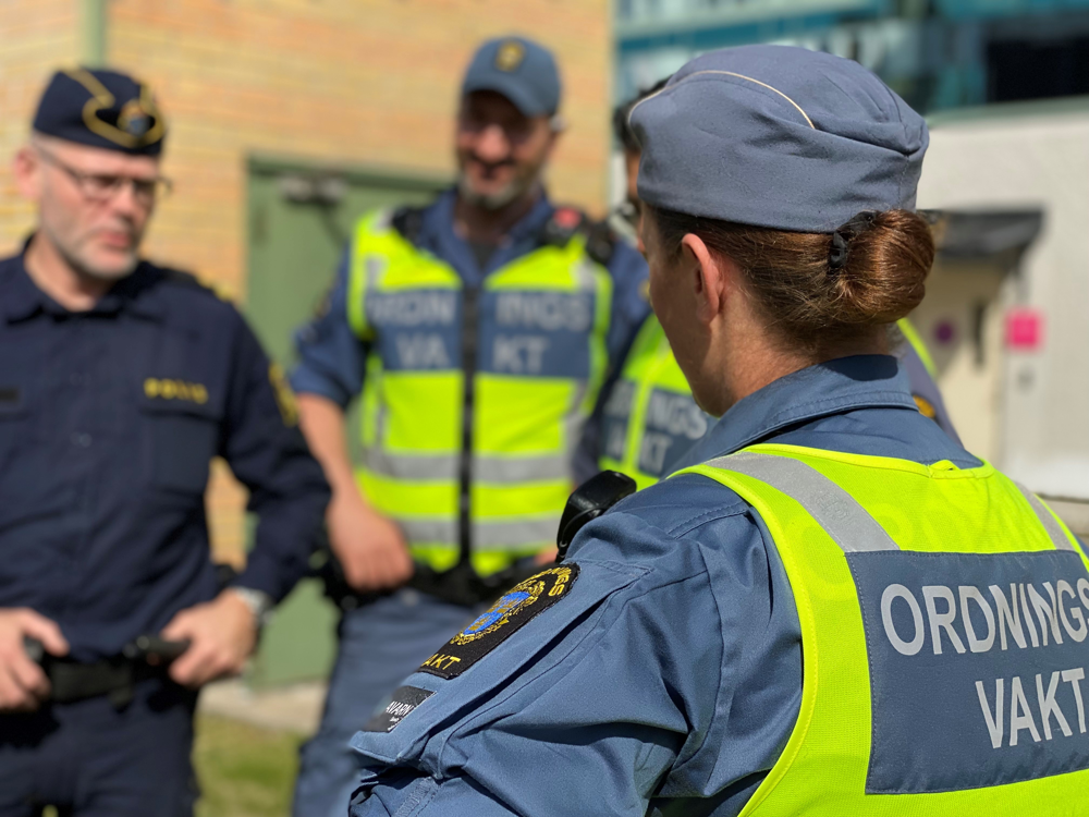 Två ordningsvakter i gula reflexvästar med texten "ordningsvakt" på ryggen samtalar med en polis.