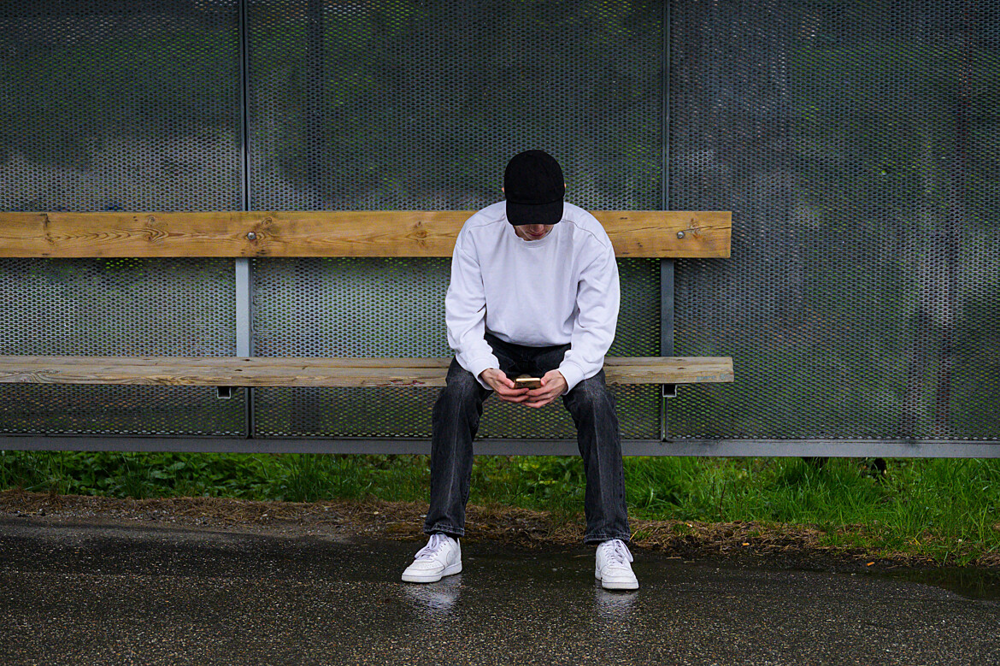 Ung person med keps sitter i en busskur med en mobiltelefon som hen tittar på.