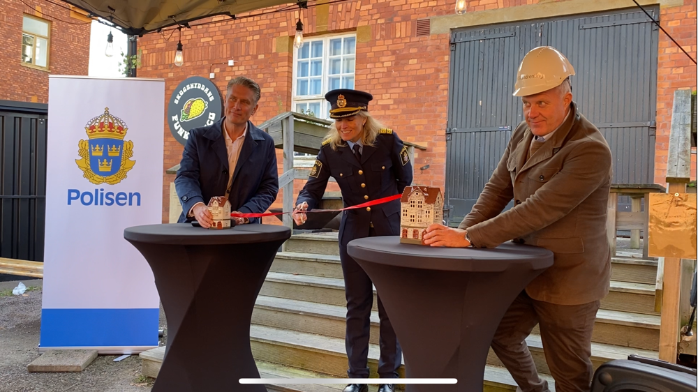 Polisområdeschefen markerar byggstart genom att klippa ett rött band som är knutet mellan två hus, ett stort och ett litet hus.