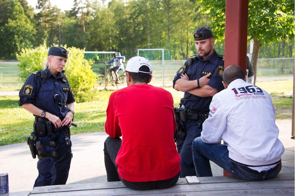 Två polisen diskuterar med ungdomar som sitter på en bänk.