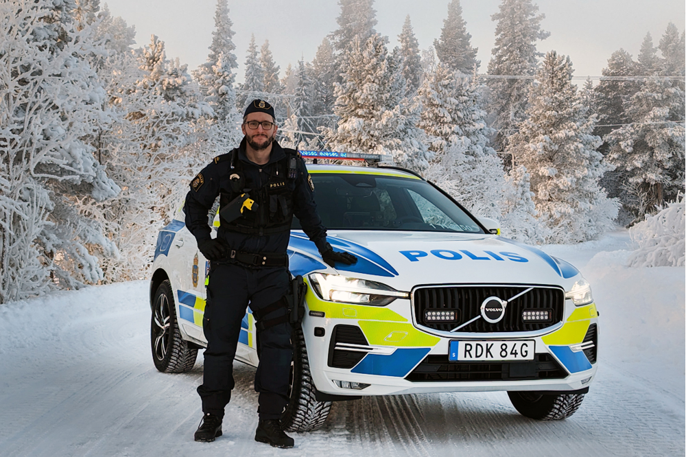 En polis står framför en polisbil. Det är vinter och snö på vägen och träden.