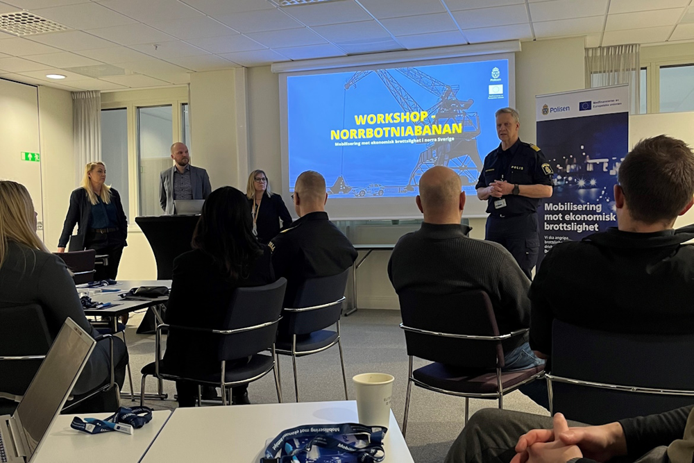 Fyra personer står uppradade framför ett antal personer som sitter ner på stolar med ryggarna vända mot kameran. Blickarna är vända mot den man som står längst till höger iklädd polisuniform. I bakgrunden visas ett bildspel med texten "Workshop - Norrbotniabanan. Mobilisering mot ekonomisk brottslighet i norra Sverige."