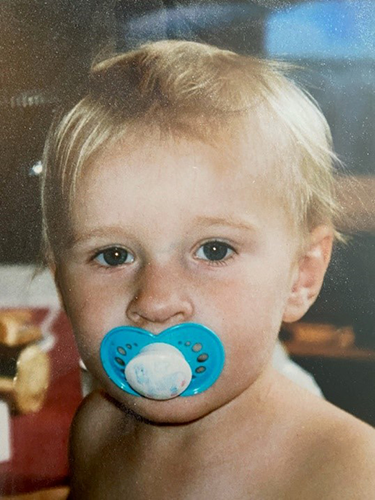 Litet barn med blont hår och en blå napp i munnen.