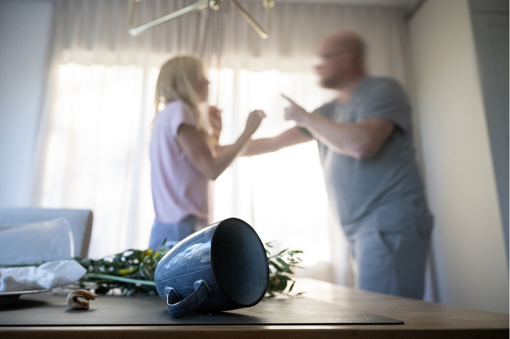 Man tar i en kvinna på ett aggressivt sätt, en temugg ligger vält på ett bord i förgrunden.