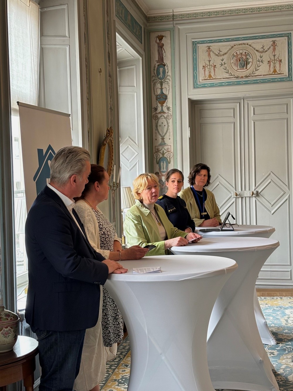 Fem personer, fyra kvinnor och en man. Står i en stor sal. Har höga bord framför sig. 