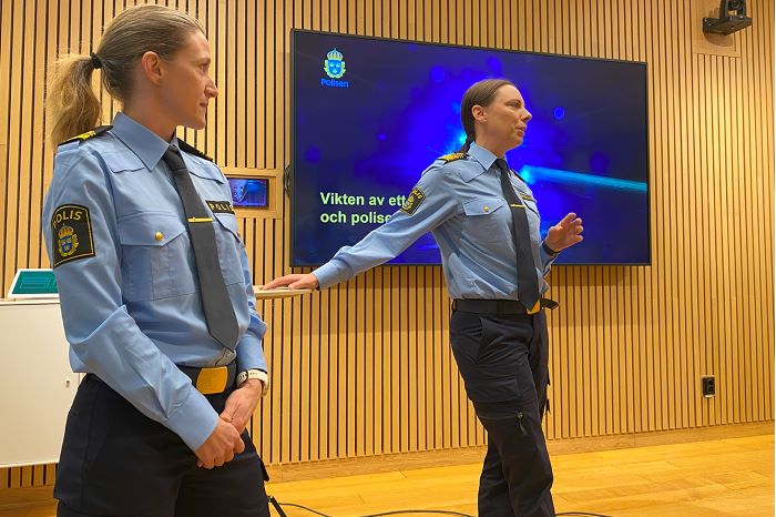 Två kvinnliga poliser framför stor bildskärm i föreläsningsrum.