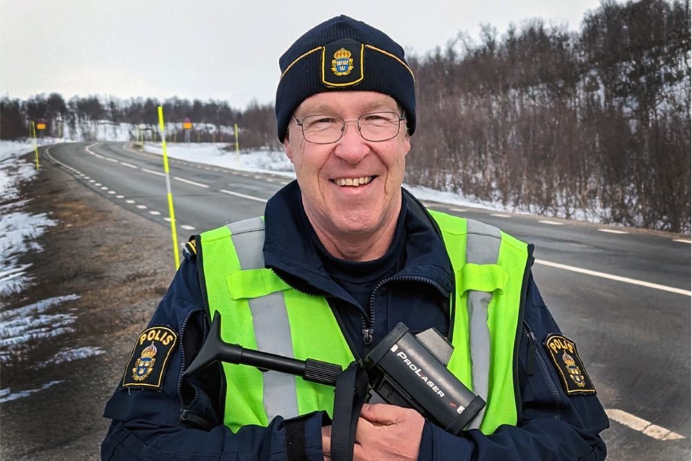 Polisman står med  ett instrument för att mäta hastighetskontroll och ler mot kameran.