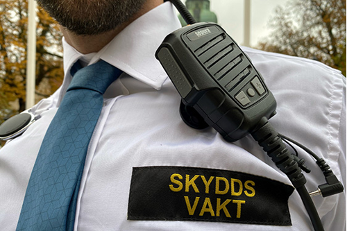 Närbild av skyddsvakt i vit skjorta med emblem med texten  "skyddsvakt". En kommunikationsradio hänger ovanför emblemet.