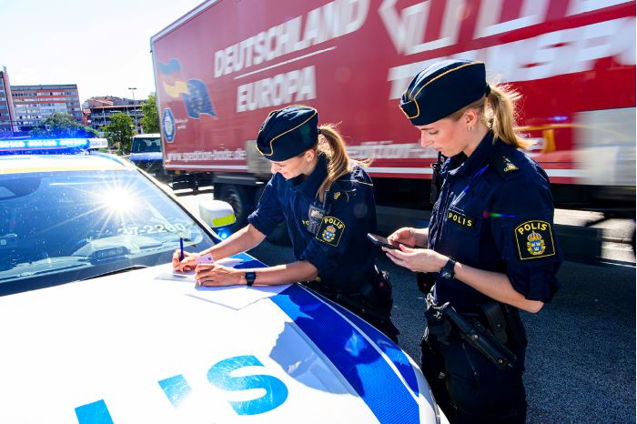 Två poliser står vid polisbilens motorhuv och skriver något. I bakgrunden syns en lastbil.
