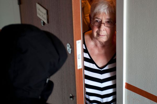 Någon med svart huva står utanför dörren när äldre kvinna öppnar och kikar ut.