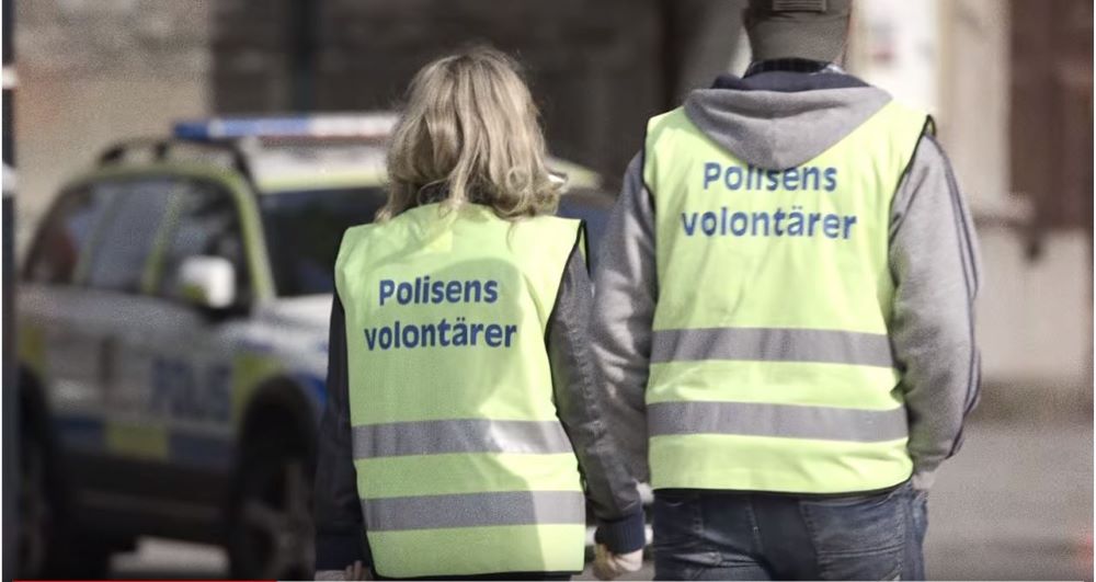 Två personer med reflexvästar med texten "Polisens volontärer" går från kameran.