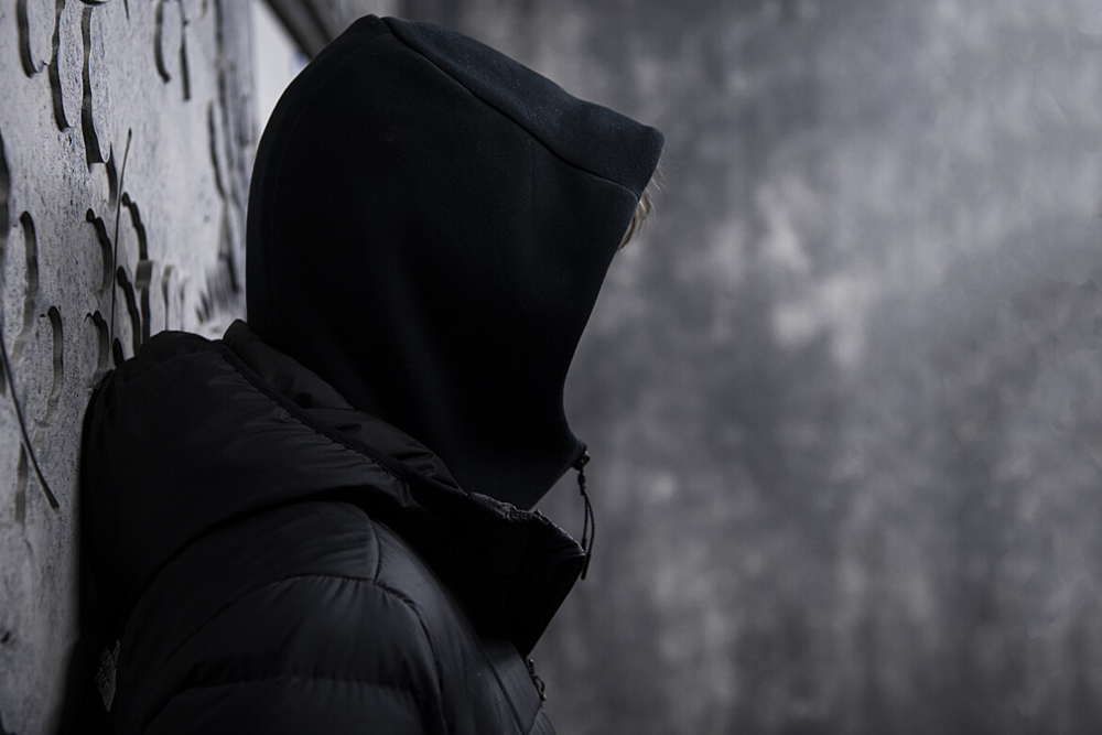 En ung person med huvtröja, där huvan täcker ansiktet, står i profil lutad mot en stenvägg. Foto.