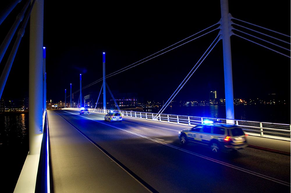 Polisbilar kör på Munksjöbron med påslaget blåljus på kvällen när bron är upplyst med blå belysning.
