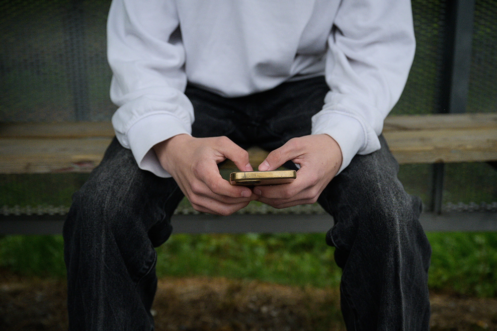 Ungdom, kille, som sitter på en bänk och tittar på sin mobiltelefon.