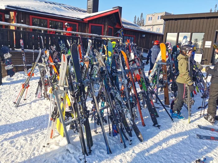 Skidor står uppradade i skidställ framför en trästuga i en skidbacke.