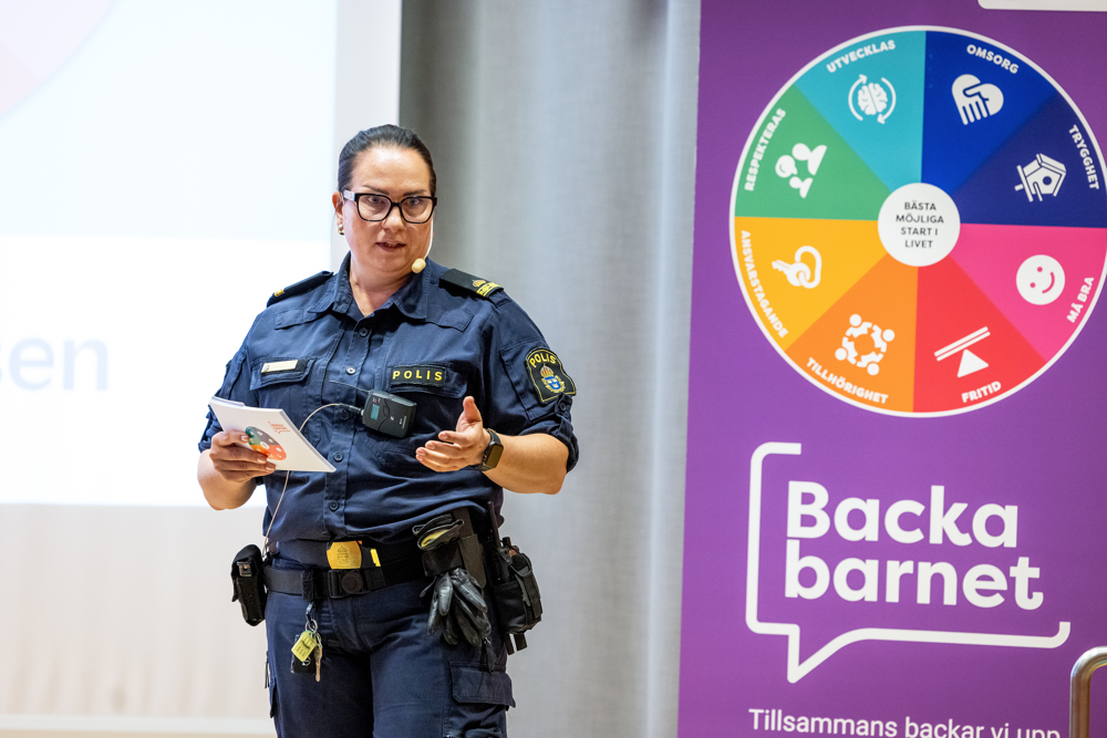 En polis föreläser på en scen bredvid en roll-up som det står Backa barnet på.