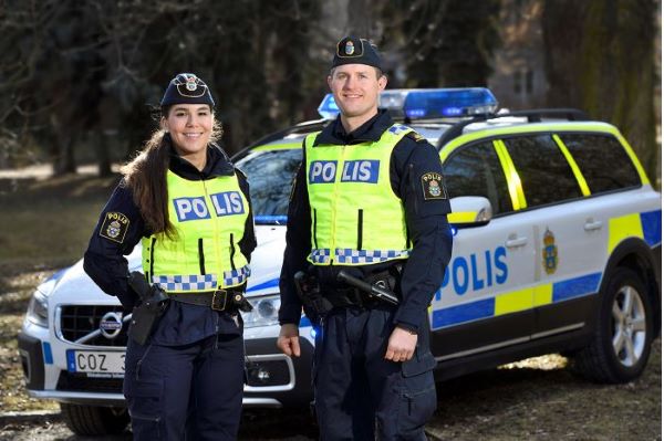 Två poliser i uniform, en kvinna till vänster och en man till höger, står utomhus framför en polisbil. De ler in i kameran.