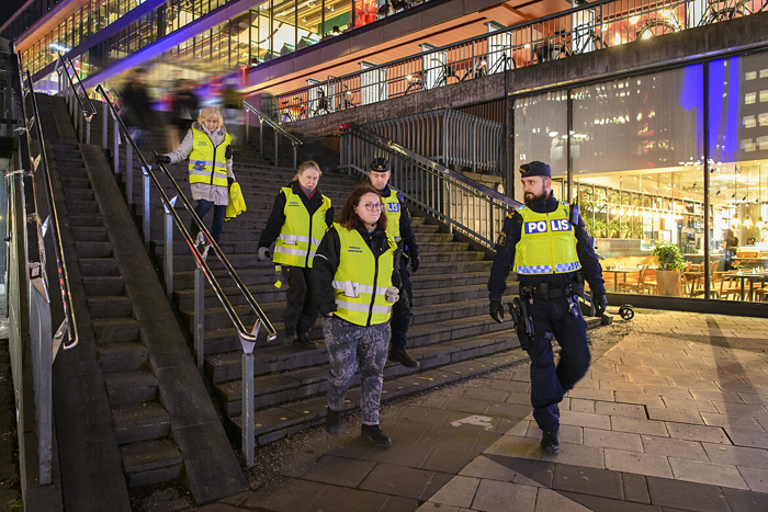 Foto på tre polisvolontärer i varselväst där det står "Polisens volontärer" som går ner för trappan vid plattan i centrala Stockholm tillsammans med två poliser i uniform.