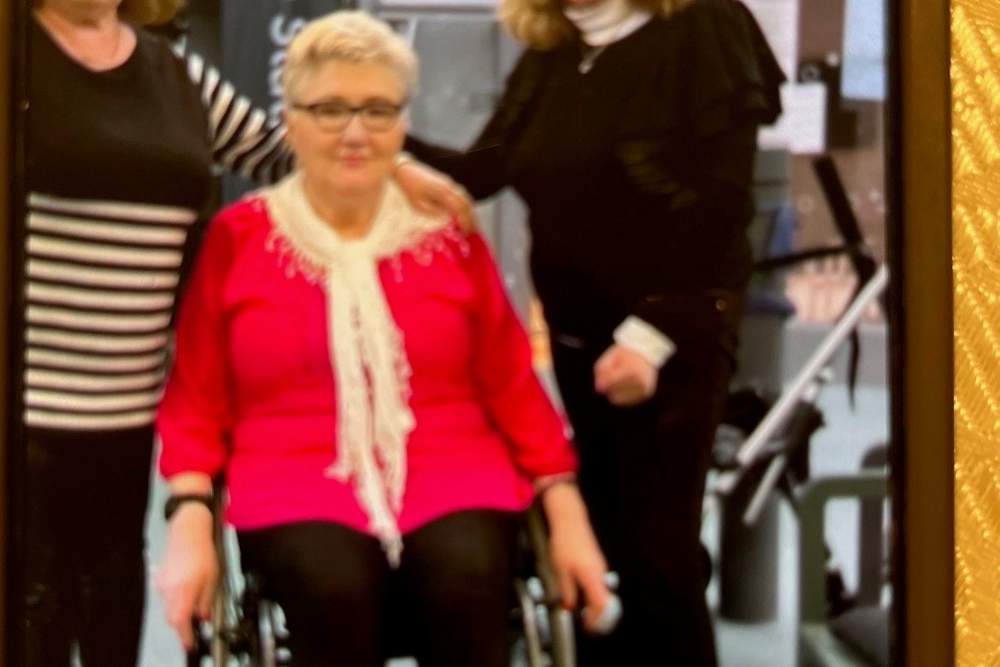 Blid på en kvinna med kort ljust hår, glasögon med svart ram som sitter i en rullstol. På bilden har kvinnan på sig en röd långärmad blus, en vit sjal och svarta byxor.