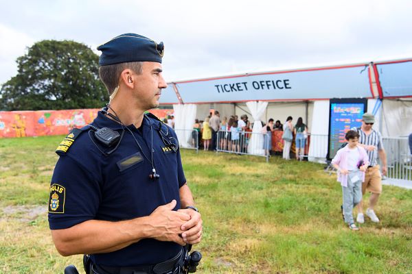 A police officer in uniform is standing on a big lawn looking at people lining up at a ticket office.