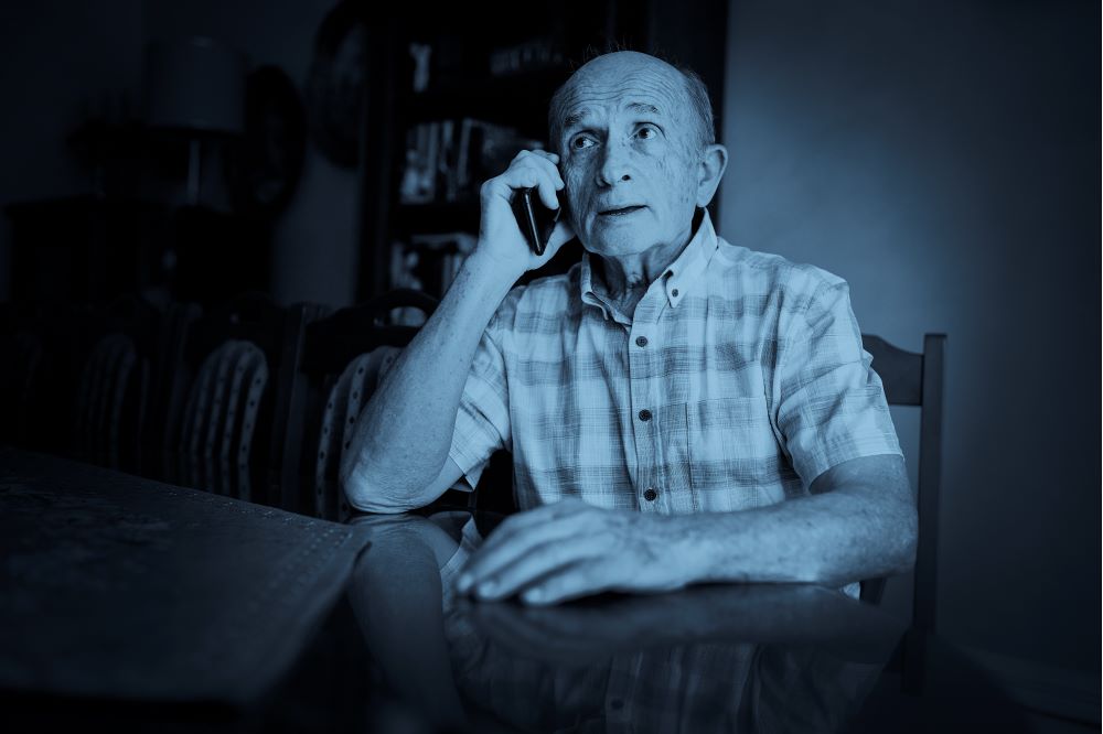 Äldre man med rutig skjorta sitter vid ett bord och pratar i mobiltelefon.